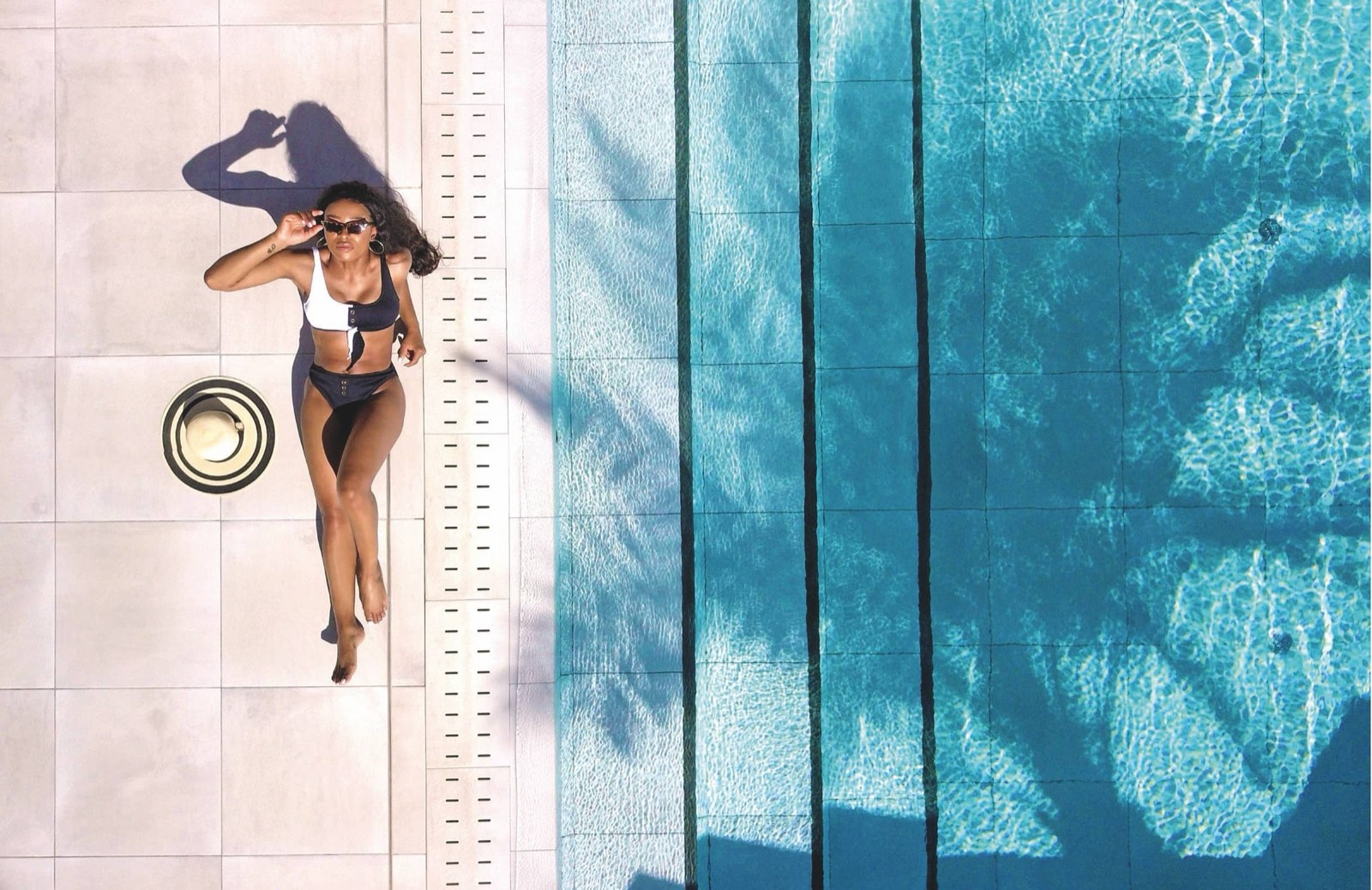 Aerial view of a woman lying poolside in a black and white swimsuit beside a straw hat
