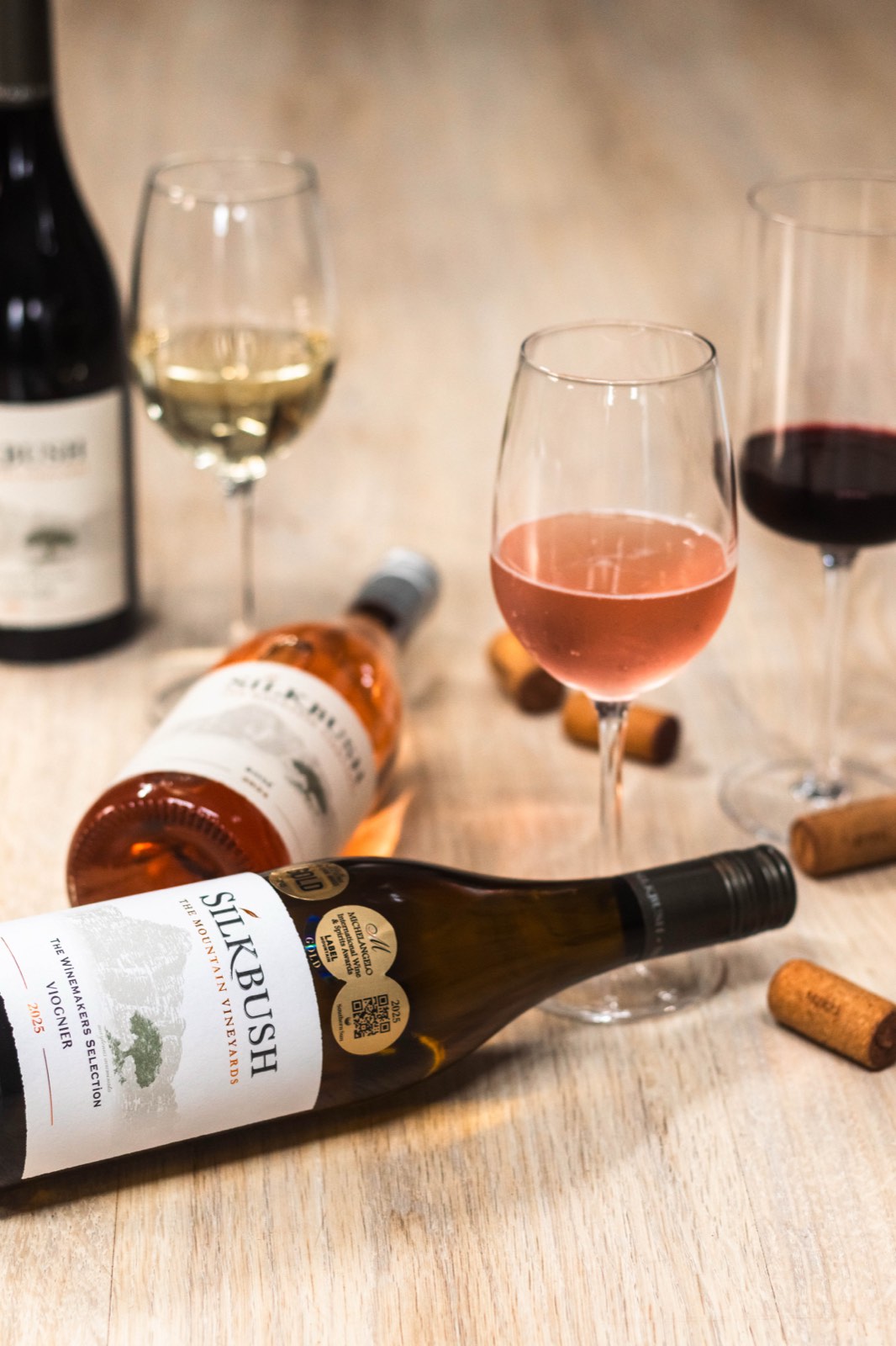 Silkbush wine bottles and glasses of red, rosé and white wine styled on a wooden table with scattered corks